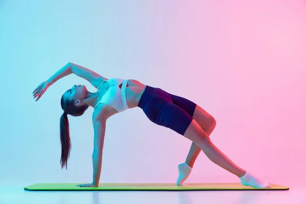 年轻女运动健将的肖像 在霓虹灯下做伸展运动 在深蓝色粉红的背景上隔离 运动的概念 身体护理 — 图库照片