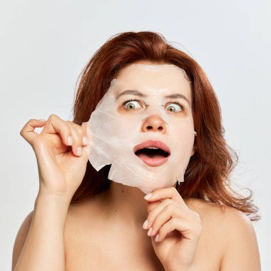 Güzel, kızıl saçlı, cildine özen gösteren, nemlendirici ve canlandırıcı maskeyi beyaz arka planda çıkartan bir kadın. Güzellik, yüz ve cilt bakımı, kozmetoloji ve doğal kozmetik kavramı