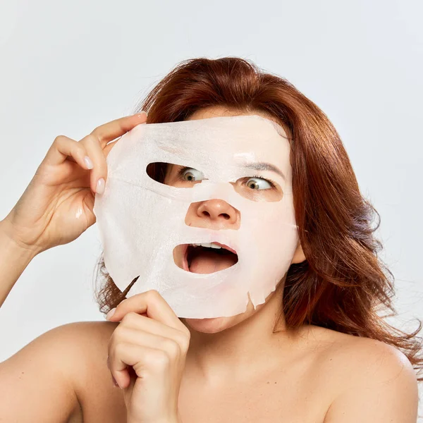 Emotive red-haired woman taking care after skin, taking off moisturizing and revitalizing mask isolated on white background. Concept of beauty, face and skin care, cosmetology, natural cosmetics, spa