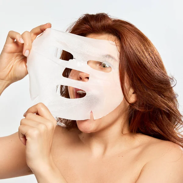 Red-haired woman emotionally taking care after skin, taking off moisturizing and revitalizing mask isolated on white background. Concept of beauty, face and skin care, cosmetology, natural cosmetics