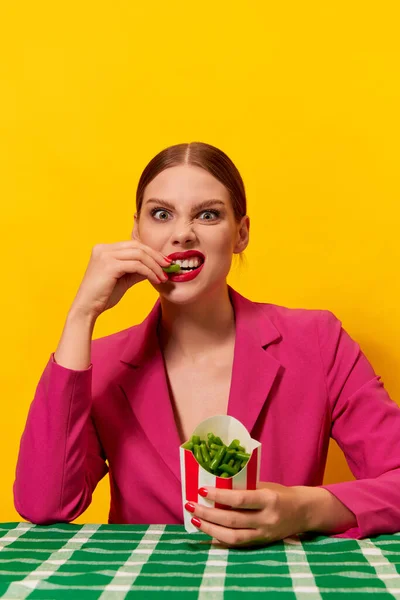 明るいピンクのジャケットの感情的な若い女性は 黄色の背景にフライドポテトパッケージから緑豆を食べています 健康的な食事 フードポップアート写真 補完的な色 テキストのコピースペース — ストック写真