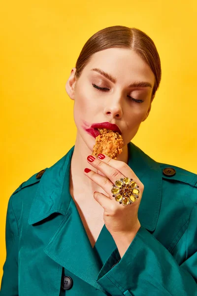 Young woman in green coat with red lipstick smudge, eating fried chicken, nuggets over yellow background. Junk food lover. Food pop art photography. Complementary colors. Copy space for ad, text