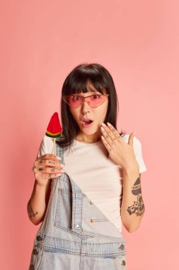 Kot tulumlu, elinde lolipopla pembe arka planda poz veren şık bir kız. Etkileyici görünüş. Gençlik kavramı, güzellik, moda, yaşam tarzı, duygular, yüz ifadesi. Ad