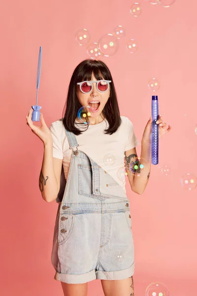 Young stylish girl in denim overalls posing with bubble blower isolated on pink background. Happy, joyful mood. Concept of youth, beauty, fashion, lifestyle, emotions, facial expression. Ad