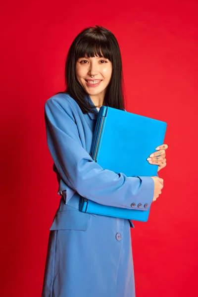 Portrait of young girl in stylish blue costume posing with laptop isolated over red background. Trainee, employee, worker. Concept of youth, beauty, fashion, lifestyle, emotions, facial expression. Ad
