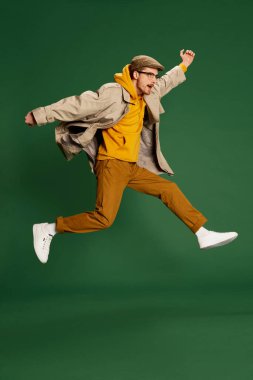 Yeşil arka planda izole bir şekilde poz veren modaya uygun sonbahar kıyafetleri giyen şık genç bir adamın portresi. Şık aksesuarlar. Gençlik kavramı, yaşam tarzı, gündelik moda, duygular, yüz ifadesi. Ad