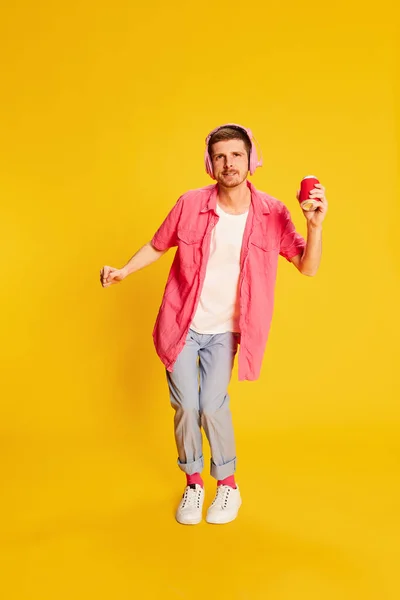 Portrait of young man in pink shirt posing, dancing in headphones isolated over vivid yellow background. Relaxed. Concept of youth, lifestyle, music, casual fashion, emotions, facial expression. Ad