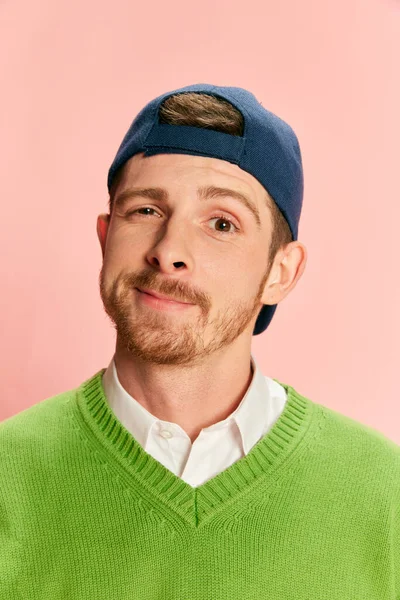 Portrait of young man in cap posing in green sweater and shirt isolated over pink background. Concept of retro fashion, sport, 90s, lifestyle, youth, emotions. facial expression. Ad