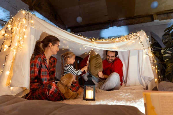 Happy family, mother, father and daughter playing inside hut with lights, tent in room in the evening. Pillows fight. Concept of fantasy, childhood, family, leisure time, love, care, home.