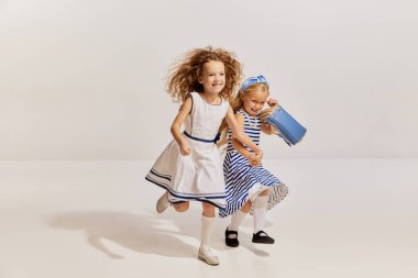 İki küçük kızın portresi, şirin elbiseli çocuklar neşeli bir şekilde koşuyorlar, gri arka planda poz veriyorlar. Mutluyum. Çocukluk kavramı, arkadaşlık, eğlence, yaşam tarzı, moda, eski tarz, duygular