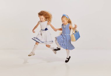 İki küçük kızın portresi, şirin elbiseli çocuklar poz veriyor, koşuyor, gri arka planda zıplıyor. Oyuncu çocuklar. Çocukluk kavramı, arkadaşlık, eğlence, yaşam tarzı, moda, eski tarz, duygular