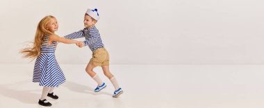 Çocuklar, erkek ve kız deniz tarzı kıyafetler giyip gri arka planda çizgili yelek giyiyorlar. El ele tutuşup dönüyorlar. Çocukluk kavramı, arkadaşlık, eğlence, yaşam tarzı, moda, eski tarz, duygular