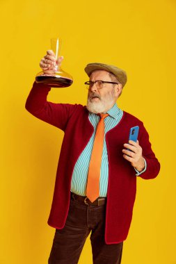 Klasik giyinmiş, gözlüklü ve şapkalı son sınıf öğrencisinin portresi. Canlı sarı arka planda şarap sürahisine bakıyor. Şarap uzmanı. Duyguların kavramı, yüz ifadesi, yaşam tarzı, modern moda.