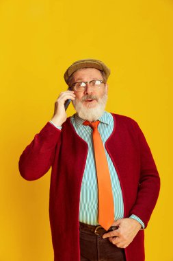 Klasik elbiseli, gözlüklü ve şapkalı neşeli son sınıf öğrencisinin portresi telefonda canlı sarı arka planda konuşuyor. Duyguların kavramı, yüz ifadesi, yaşam tarzı, modern moda.