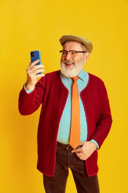 Klasik kıyafetler, gözlük ve şapka takmış yaşlı bir adamın portresi telefonda canlı sarı arka planda görüntülü konuşma. Duyguların kavramı, yüz ifadesi, yaşam tarzı, modern moda.