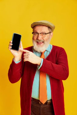 Klasik elbiseli, gözlüklü ve şapkalı mutlu son sınıf öğrencisinin portresi canlı sarı arka planda telefon ekranını işaret ediyor. Duyguların kavramı, yüz ifadesi, yaşam tarzı, modern moda.