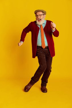 Klasik elbiseli, gözlüklü ve şapkalı neşeli son sınıf öğrencisinin portresi, parlak sarı arka planda dans ediyor. Duyguların kavramı, yüz ifadesi, yaşam tarzı, modern moda.