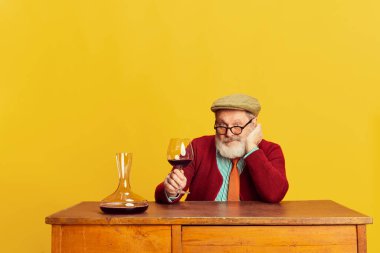 Klasik elbiseli, gözlüklü ve şapkalı son sınıf öğrencisinin portresi canlı sarı arka planda şarapla poz veriyor. Profesör. Duyguların kavramı, yüz ifadesi, yaşam tarzı, modern moda.