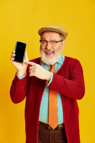 Klasik elbiseli, gözlüklü ve şapkalı mutlu son sınıf öğrencisinin portresi canlı sarı arka planda telefon ekranını işaret ediyor. Duyguların kavramı, yüz ifadesi, yaşam tarzı, modern moda.