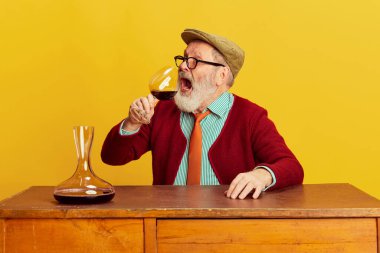 Klasik giyinmiş, gözlüklü, şapkalı, parlak sarı arka planda şarap içen yaşlı bir adamın portresi. Duyguların kavramı, yüz ifadesi, yaşam tarzı, modern moda.