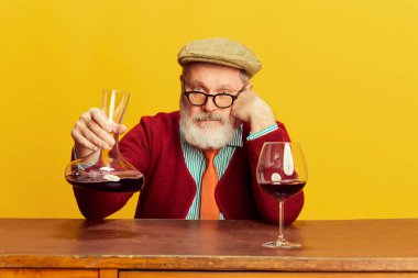 Portrait of senior man in classical clothes, glasses and cap posing with red wine over vivid yellow background. Concept of emotions, facial expression, lifestyle, modern fashion clipart