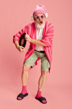 Şık bir son sınıf öğrencisi, şık parlak kıyafetli yaşlı bir adam örgü şapkalı, güneş gözlüklü ve kulaklıklı pembe arka planda plakla poz veriyor. Duyguların kavramı, yüz ifadesi, yaşam tarzı, modern moda.