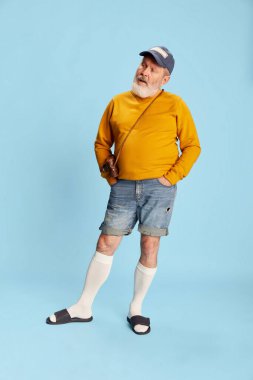 Mavi arka planda kasketli ve klasik fotoğraf makineli sıradan giyinmiş yaşlı sakallı bir adamın portresi. Gezgin. Duyguların kavramı, yüz ifadesi, yaşam tarzı, modern moda.