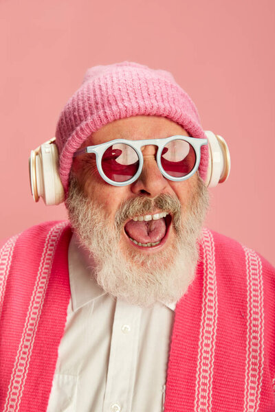 Portrait of stylish senior, old man in stylish bright outfit with knitted hat and sunglasses posing in headphones on pink background. Concept of emotions, facial expression, lifestyle, modern fashion