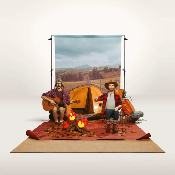 Two men recreating camping activity over grey background with nature wallpaper. Sitting near fire and playing guitar. Concept of travelling, active lifestyle, friendship, leisure activity, relaxation