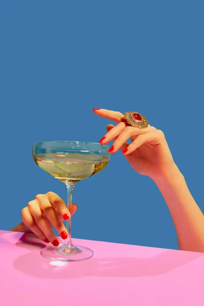 Female hands holding glass with champagne over blue pink background. Festive celebration. Food aesthetics. Concept of holiday, party, drink. Complementary colors. Copy space for ad. Pop art