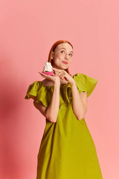 Portrait of young beautiful girl in cute dress posing with cupcake over pink background. Cute, dreamy look. Concept of youth, beauty, fashion, lifestyle, emotions, facial expression. Ad