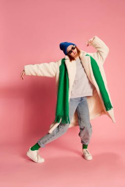 Güneş gözlüklü, mavi şapkalı, yeşil atkı ve pembe arka planda poz veren kürklü genç kız. Sıradan. Gençlik kavramı, güzellik, kış modası, yaşam tarzı, duygular, yüz ifadesi. Ad