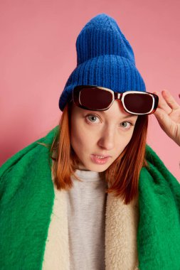 Güneş gözlüklü, mavi şapkalı, yeşil atkı ve pembe arka planda poz veren kürklü genç kız. Sorgulayıcı bakış. Gençlik kavramı, güzellik, kış modası, yaşam tarzı, duygular, yüz ifadesi. Ad