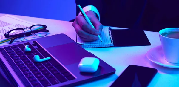 Business person making notes. Laptop keyboard, headphones, glasses and phone on table. Working space in neon light. Concept of business, modern technologies, occupation, career development
