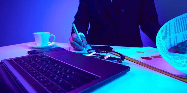 Working desk with laptop, notebook, coffee, phone in neon colored light. Making notes, writing down tasks. Concept of business, modern technologies, occupation, career development