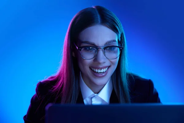 Happy woman in stylish official suit working on laptop over blue background in neon light. Success and motivation. Concept of business, modern technologies, occupation, career development