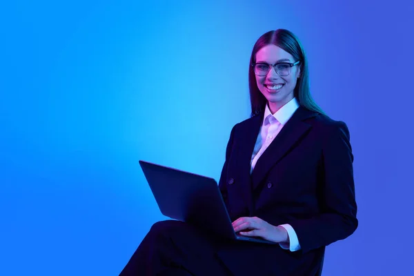 Woman in stylish official suit working on laptop over blue background in neon light. Successful business woman. Concept of business, modern technologies, occupation, career development