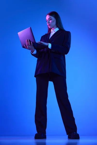 Woman in stylish official suit working on laptop over blue background in neon light. Concentrated business woman. Concept of business, modern technologies, occupation, career development