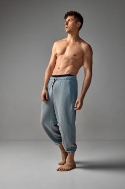Gri stüdyo arka planında pantolonla poz veren yakışıklı, üstsüz, olgun bir adam. Kas gevşetici vücut şekli. Sağlıklılık. Erkeklerin sağlık ve güzellik anlayışı, vücut ve cilt bakımı, fitness. Vücut sanatı