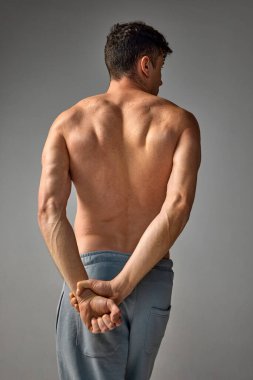 Arkadan rahatlama, kaslı, güçlü erkek vücudu, sırt ve eller. Tişörtsüz poz veren olgun bir adam, gri stüdyo arka planında pantolonu var. Erkeklerin sağlık ve güzellik kavramı, vücut ve cilt bakımı, sağlık.