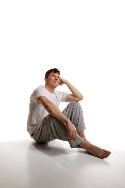 Rahat görün. Tişört ve pantolonla yerde oturan olgun gülümseyen adam beyaz stüdyo arka planında. Ev ödevi. Rahatlatıcı. Erkeklerin sağlık ve güzellik anlayışı, vücut ve cilt bakımı, fitness. Vücut sanatı