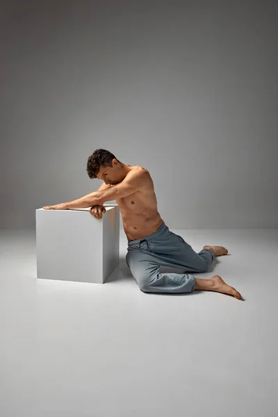 Body aesthetics. Mature handsome man sitting on floor, posing shirtless in pants over grey studio background. Concept of mens health and beauty, body and skin care, fitness. Body art