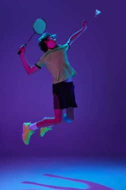 Badminton oynayan üniformalı bir gencin portresi neon ligth ile izole edilmiş mavi bir arka planda mekik horozu servis ediyor. Spor yaşam tarzı kavramı, hareket, eylem, rekabet