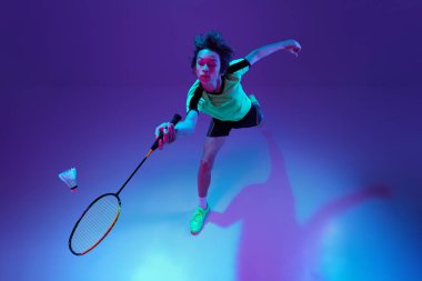 Üst Manzara. Badminton oynayan üniformalı bir gencin portresi neon ligth ile mavi mor arka planda mekik sikini iade ediyor. Spor yaşam tarzı kavramı, hareket, eylem, rekabet