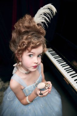 Şirin küçük bir kız, ortaçağdan kalma muhteşem elbiseli bir çocuk piyanonun başında oturmuş, duygusal olarak kameraya bakıyor. Surat asma. Tarihi yeniden yapılanma kavramı, çağların karşılaştırması, ortaçağ modası