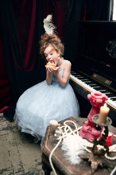 Portrait of cute little girl, child in image of medieval royal person emotionally eating birthday cake. Concept of historical remake, comparison of eras, medieval fashion, emotions, childhood