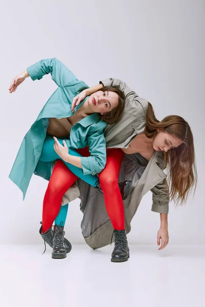 Extraordinary. Portrait of two young women in bright red and blue tights and coat posing over grey studio background. Concept of modern fashion, queer, art photography, weird people, creativity