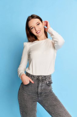 Şirin, gülümseyen. Beyaz bluzlu ve kot pantolonlu genç güzel bir kızın portresi mavi stüdyo arka planında poz veriyor. Gençlik kavramı, güzellik, moda, yaşam tarzı, duygular, yüz ifadesi. Ad