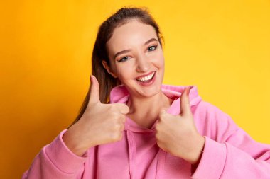 Pembe kapüşonlu güzel bir kızın portresi, sarı arka plan üzerinde onay ve pozitiflik göstergesi. Gençlik kavramı, güzellik, moda, yaşam tarzı, duygular, yüz ifadesi. Ad