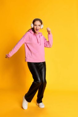 Pembe kapüşonlu genç kız portresi, kulaklıkla müzik dinliyor ve sarı stüdyo arka planında dans ediyor. Gençlik kavramı, güzellik, moda, yaşam tarzı, duygular, yüz ifadesi. Ad